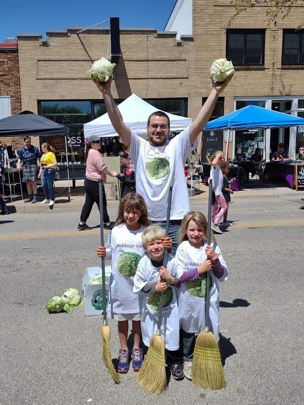 Man and three young children dressed in white t-shirts and aprons with cabbage images, holding brooms, standing on street at outdoor market or festival; man holds large leafy vegetables like cabbage above his head, kids hold brooms, others in backgro