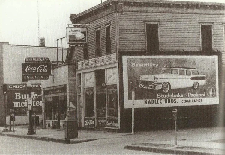 Black-and-white photo of a corner building with advertisements and signs, including a large billboard for Studebaker-Packard cars, a Coca-Cola sign, and signs for a pharmacy, magazines, and a burger restaurant.