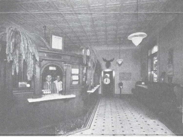 A black and white photo of a vintage hotel or inn lobby with a checkered floor, wooden counter, and hanging light fixtures. Two people stand behind the counter, and decorative items such as a mounted animal head and pendulum clock are visible on the 
