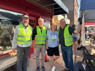 Four people wearing high visibility vests standing on a sidewalk outside a store with an 'OPEN' sign, three men and one woman smiling.