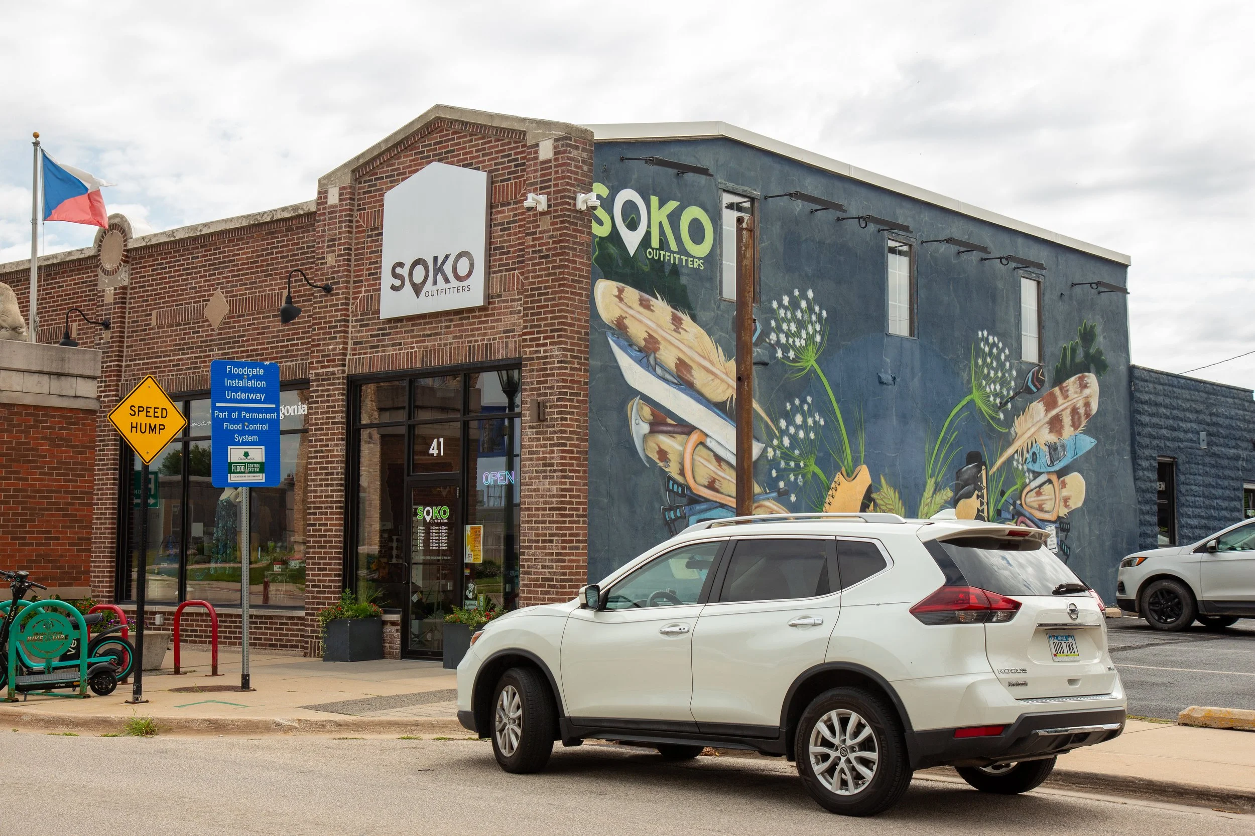 The exterior of SOKO Outfitters store in a brick building with a colorful mural on one side depicting insects and flowers. There is a sign with the store's name, a flag, parking signs, and cars parked in front.