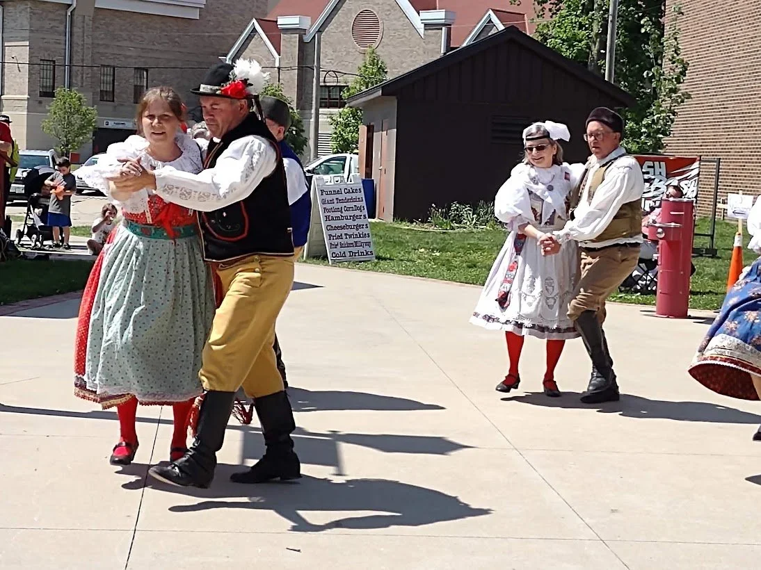 People in traditional European folk costumes dancing outdoors on a sunny day.