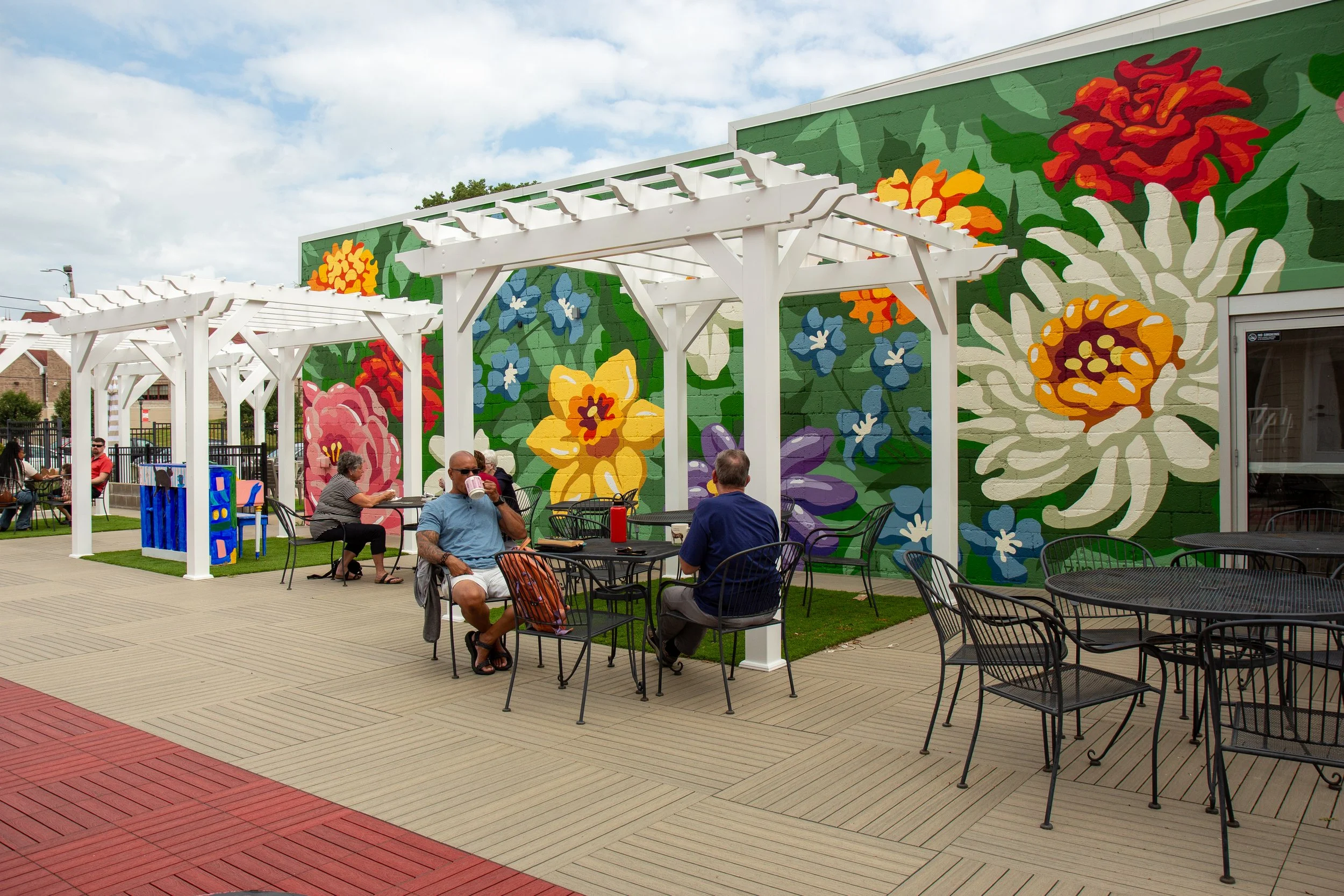 People sitting on patio furniture under white pergolas outside with colorful floral mural on green building wall in background.