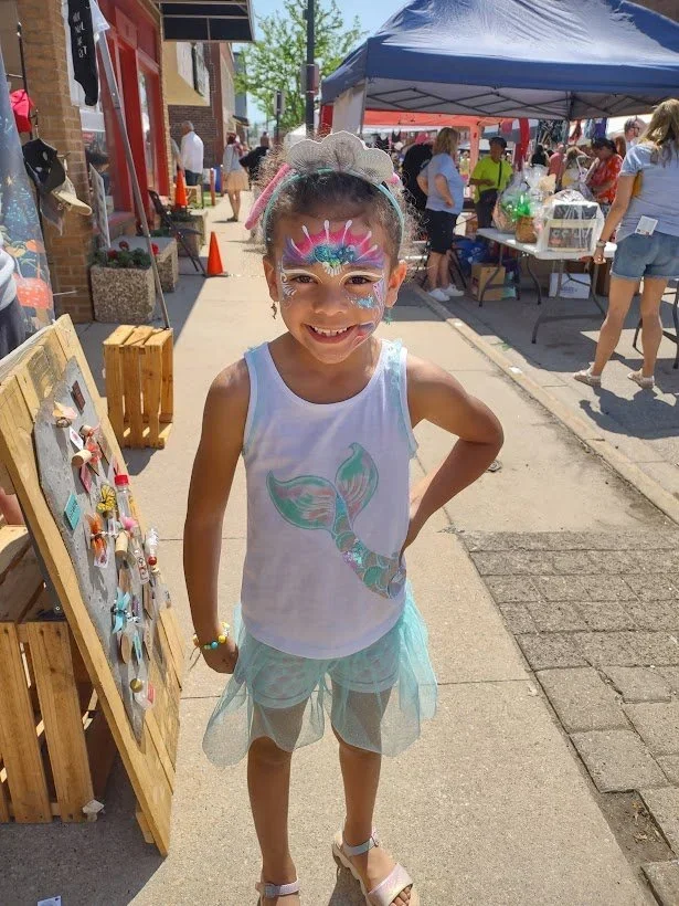Young girl with mermaid face paint at outdoor market, wearing a sleeveless top with mermaid tail design, tulle skirt, smiling.