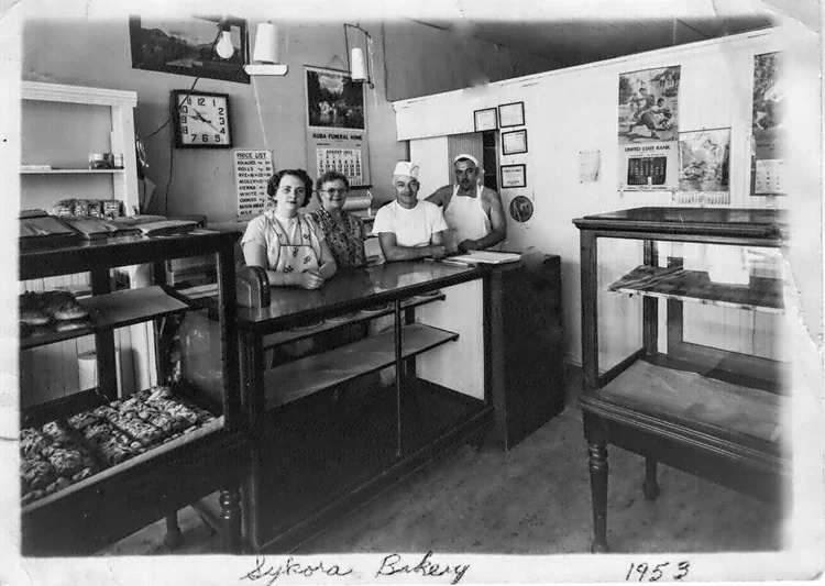 Four bakery workers standing behind a counter in a bakery shop, with display cases and baked goods, dated 1953.