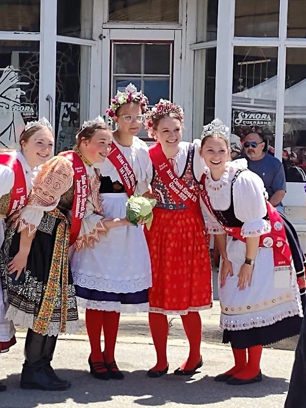 Group of young girls dressed in traditional Polish folk costumes, wearing floral crowns and sashes, standing outside a building, smiling for a photo.