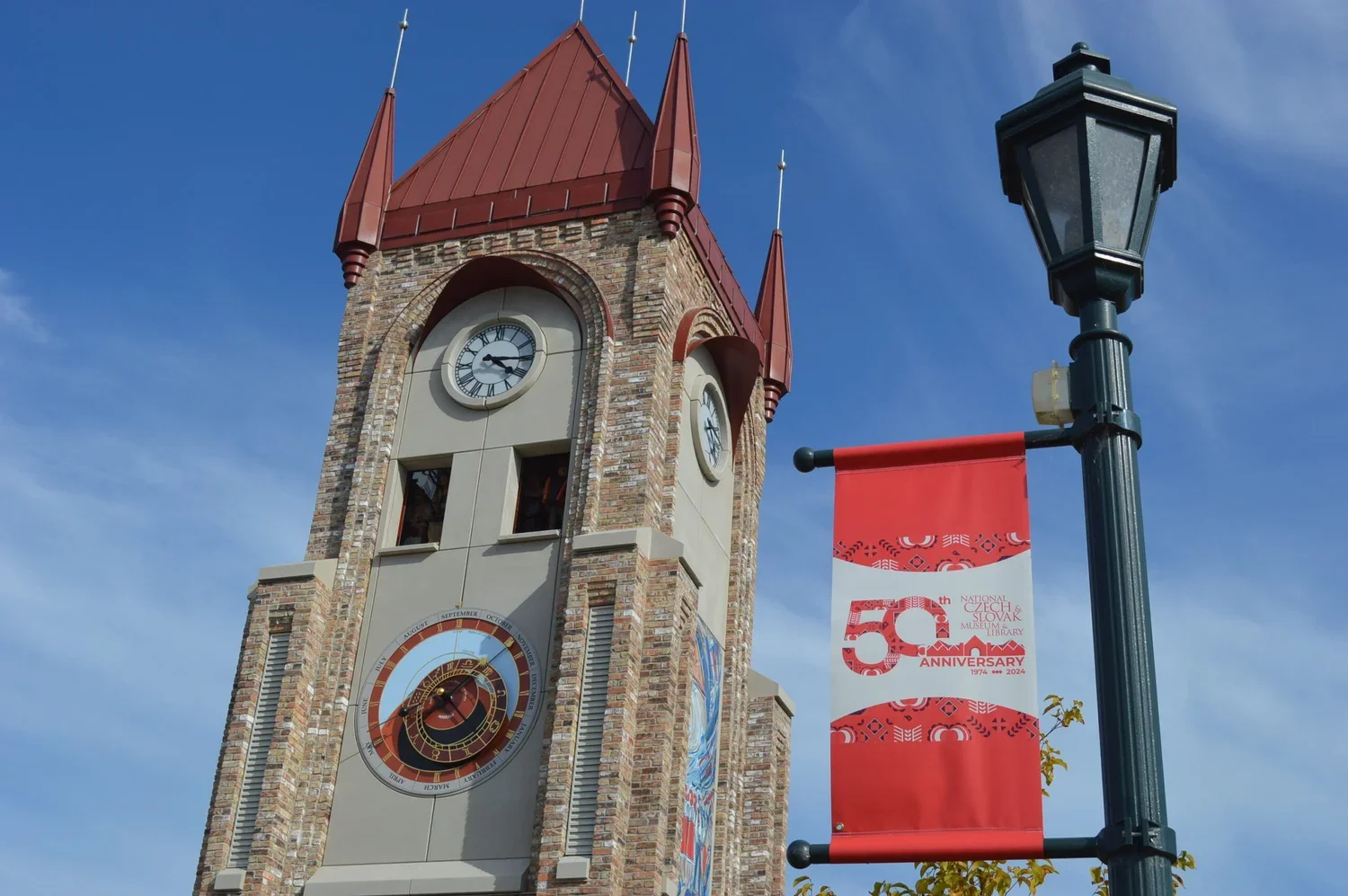 A clock tower with a red metal roof and brick facade, displaying the time 3:16, and an astronomical calendar below the clock, next to a street lamp with a banner celebrating the 50th anniversary of the Czech & Slovak Museum and Library.