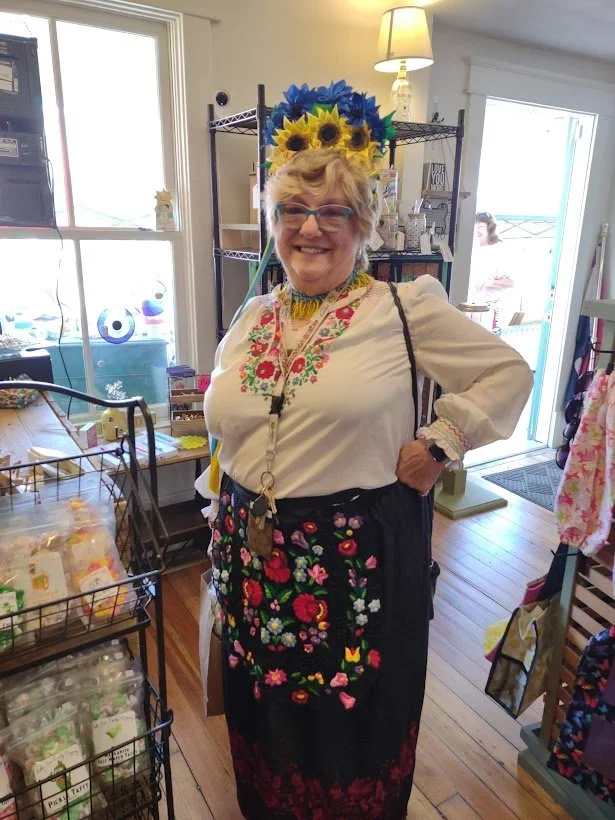 Smiling woman with short gray hair and glasses, wearing a sunflower headpiece, embroidered blouse, and a black apron with colorful floral embroidery, inside a shop.