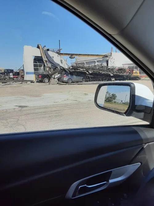 A building on its side with a collapsed roof and debris scattered, including a large metal structure or sign frame. Several vehicles, including a van and cars, are parked near the building. The photo is taken from inside a car, with the side mirror a