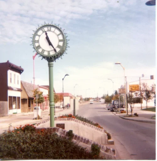 A vintage street scene featuring a large outdoor clock showing 4:50, storefronts, parked cars, streetlights, and a cloudy sky.