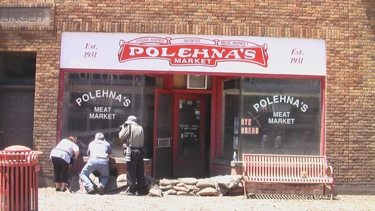 The front of Polehna's Market, a meat market established in 1931, with a large white sign featuring red lettering and logo. Three people are gathered near the window, and there is a bench and a red trash can outside.