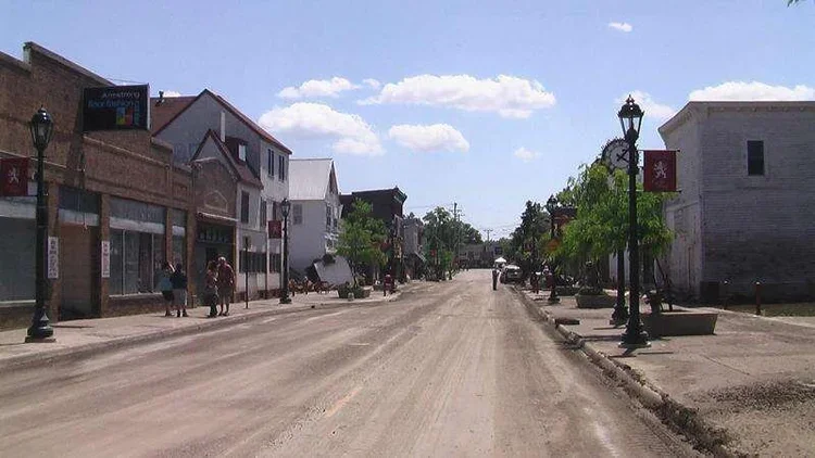 Main street in a small town with shops, a clock, street lamps, and people walking on the sidewalk under a partly cloudy sky.
