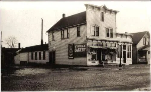 Black and white photo of a two-story bakery building with a sign that says 'Sykora Bakery'. The building has a porch and an awning in front. There are residential houses nearby.