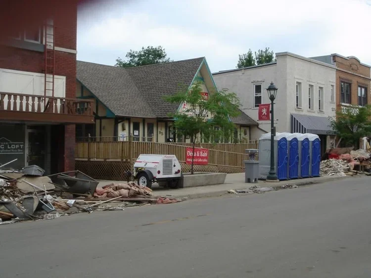 Street view with construction debris, portable toilets, and storefronts, including a building with a red sign.