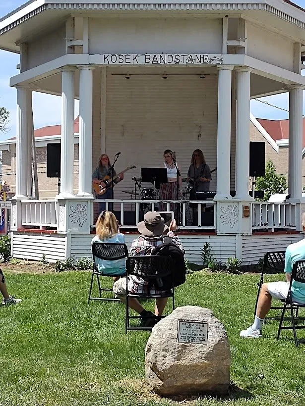 A band performing on an outdoor bandstand stage labeled "KOSEK BANDSTAND" with three musicians playing guitars and singing. Audience members are seated on chairs in front of the stage, watching the performance.