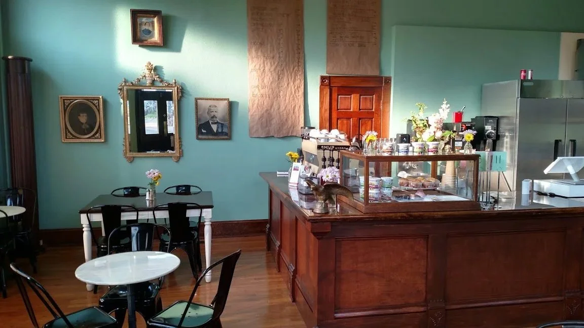 Interior of a cozy cafe with mint green walls, vintage décor, a coffee counter with glass display, and black and white tables and chairs.