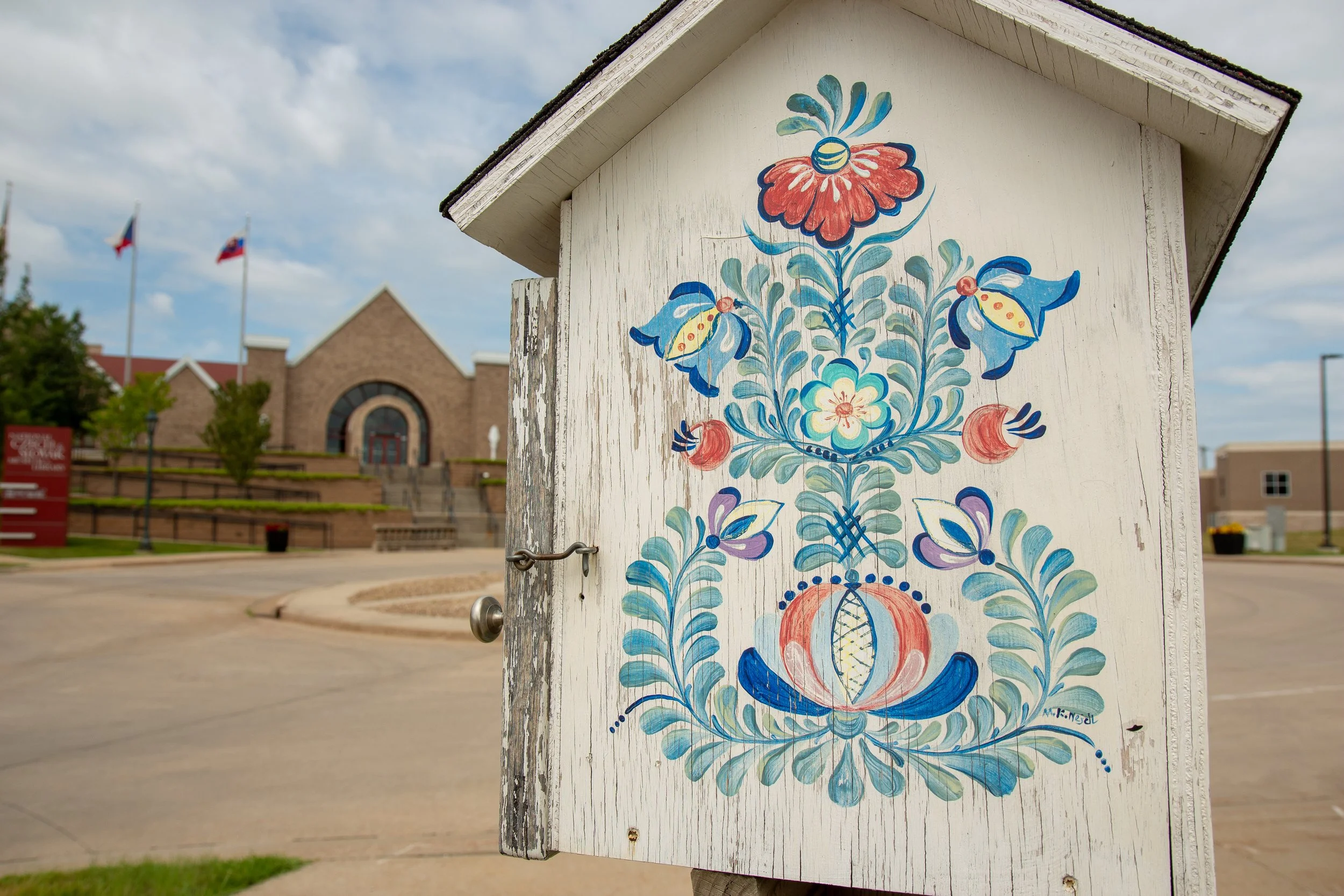A decorative painted wooden box with floral and butterfly designs on its front, located outdoors near a street with a building, flags, and cloudy sky in the background.