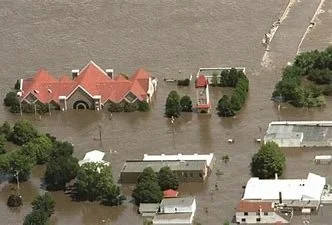 Aerial view of a flooded neighborhood with water covering streets and lawns, houses surrounded by trees, and floodwaters extending across the area.