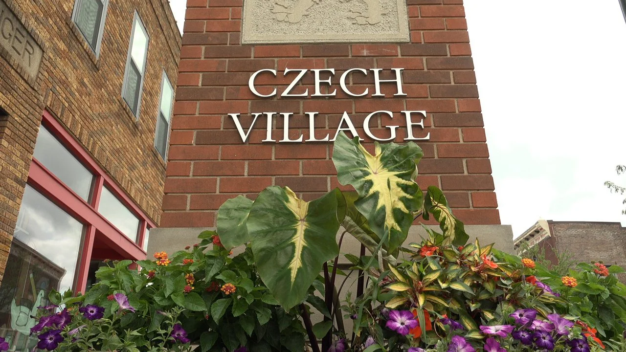 Sign that reads 'Czech Village' on a brick building with colorful flowers and green plants in the foreground.
