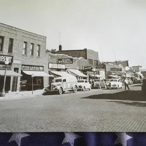 Black and white photo of small-town main street with vintage cars parked along the sidewalk, storefronts and signs for grocery store, variety store, and other businesses.