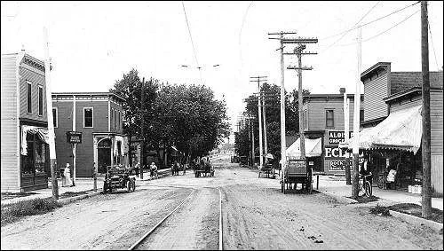 Black and white photo of a small town street with buildings, trees, utility poles, and horse-drawn carriages.