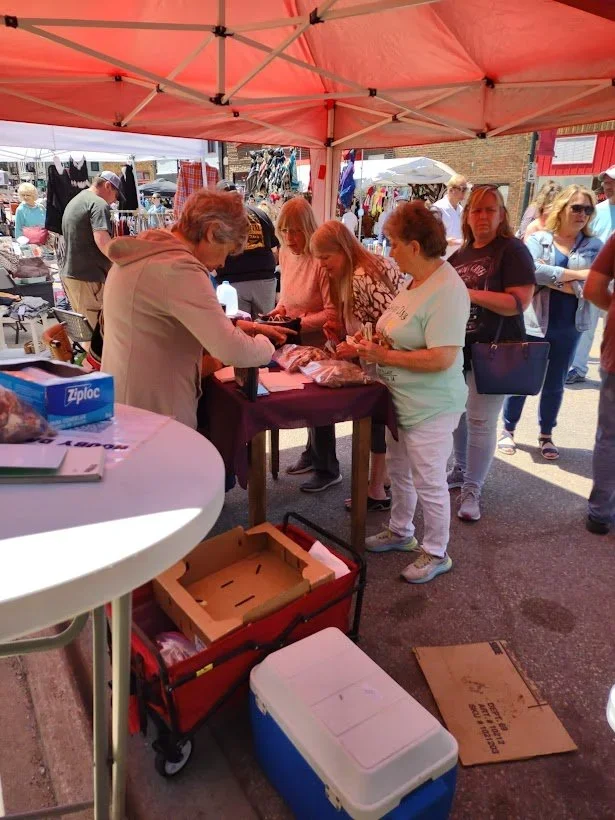 People shopping at an outdoor market stall under a red canopy, with various items on display and several individuals browsing and purchasing.