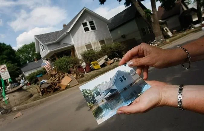 Person holding a picture of a house with floodwaters in front of the actual house showing flood damage.