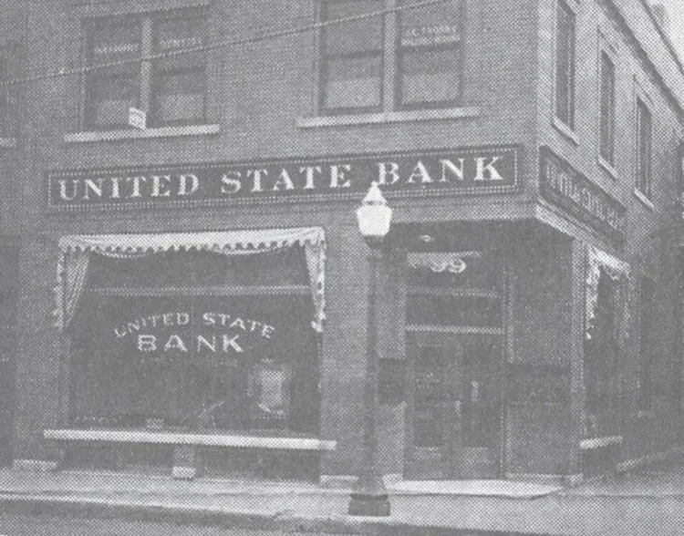 Black and white photo of the corner of a brick building with a sign reading 'United State Bank' and a window display with the same name.