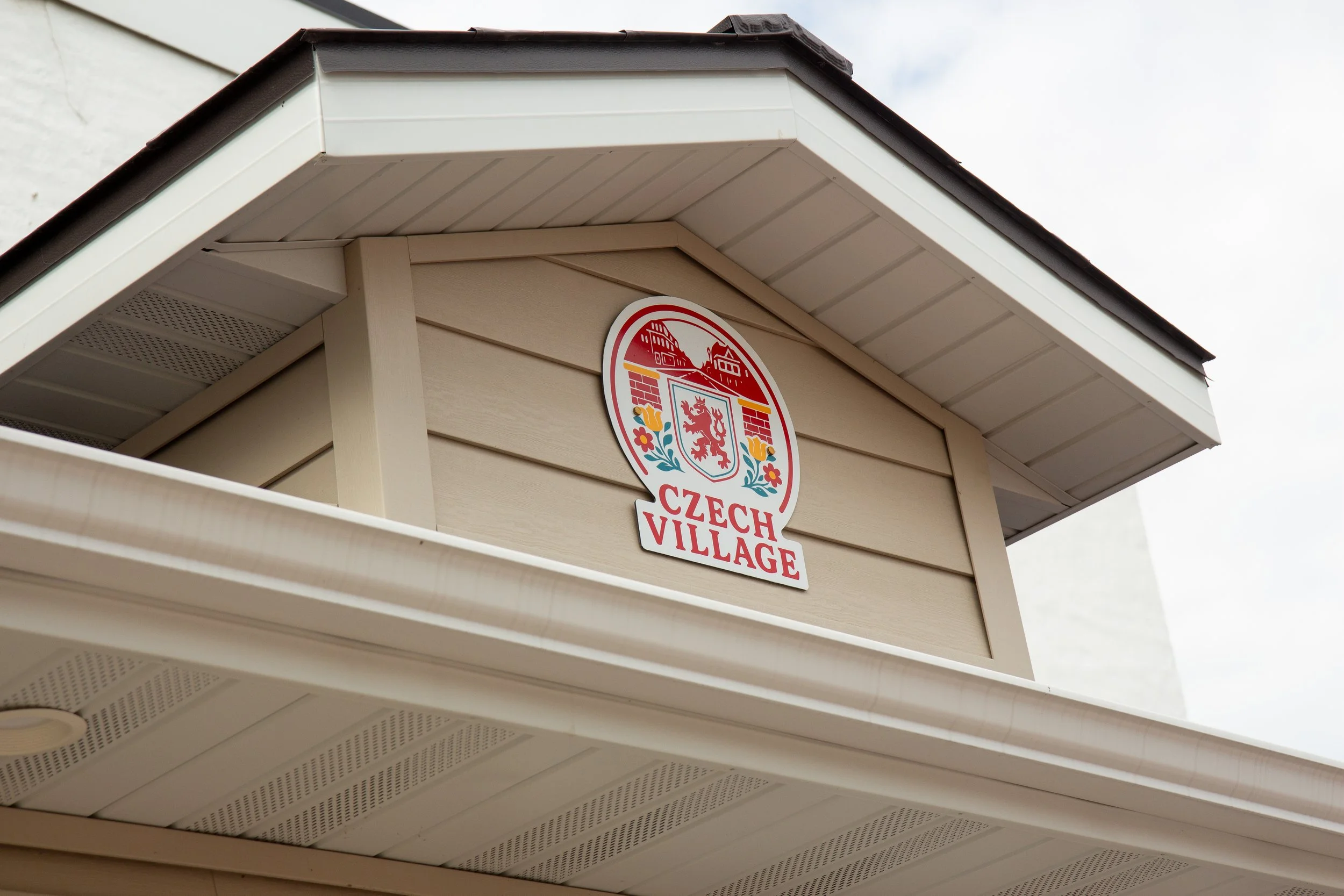 Close-up of a wooden structure with a sign that reads 'Czech Village' and a coat of arms featuring a lion, bricks, and flowers.