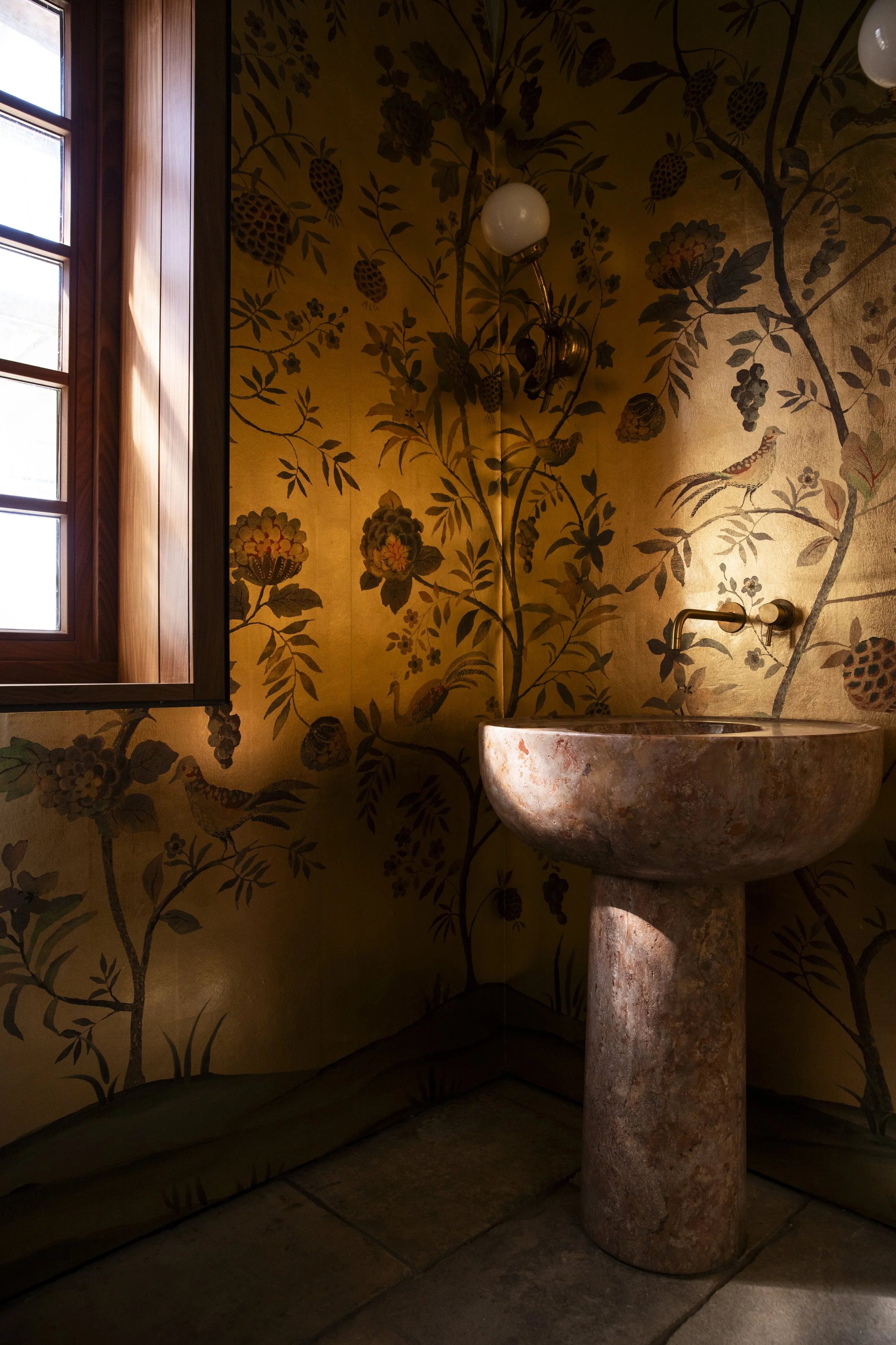 Vintage-style sink and wall lamp in a room with gold floral wallpaper and wooden window frame.