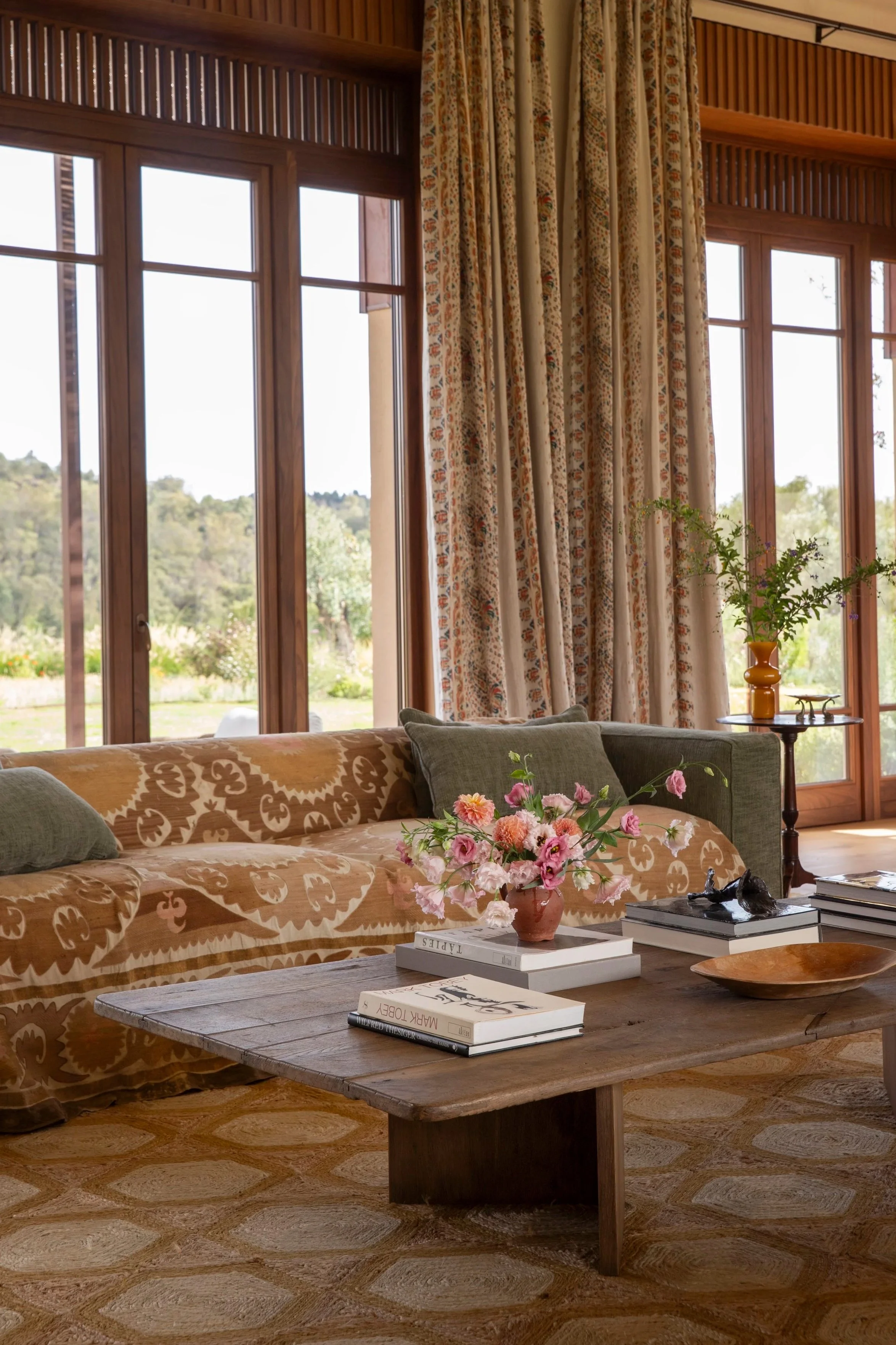 Living room with large windows, beige patterned couch with pillows, wooden coffee table with books and a flower arrangement, and patterned curtains, overlooking a green outdoor landscape.