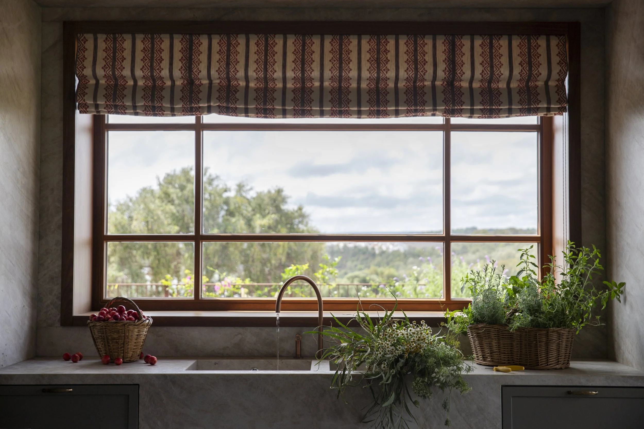 A kitchen window with decorative window valance, view of trees outside, windowsill with baskets of red tomatoes, green herbs, and kitchen sink faucet.