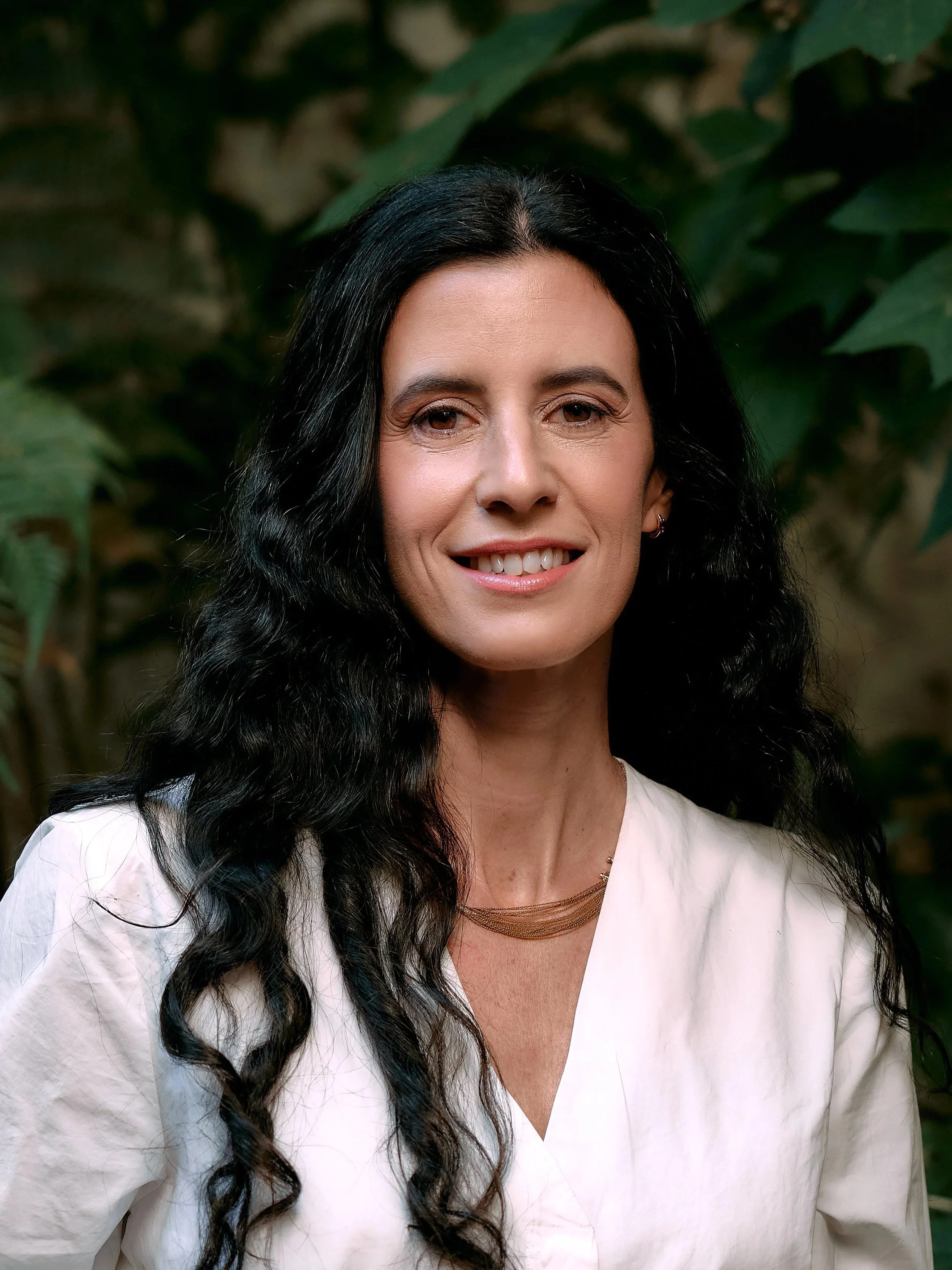 A woman with long black wavy hair, wearing a white blouse and gold jewelry, smiling, with green leafy plants in the background.