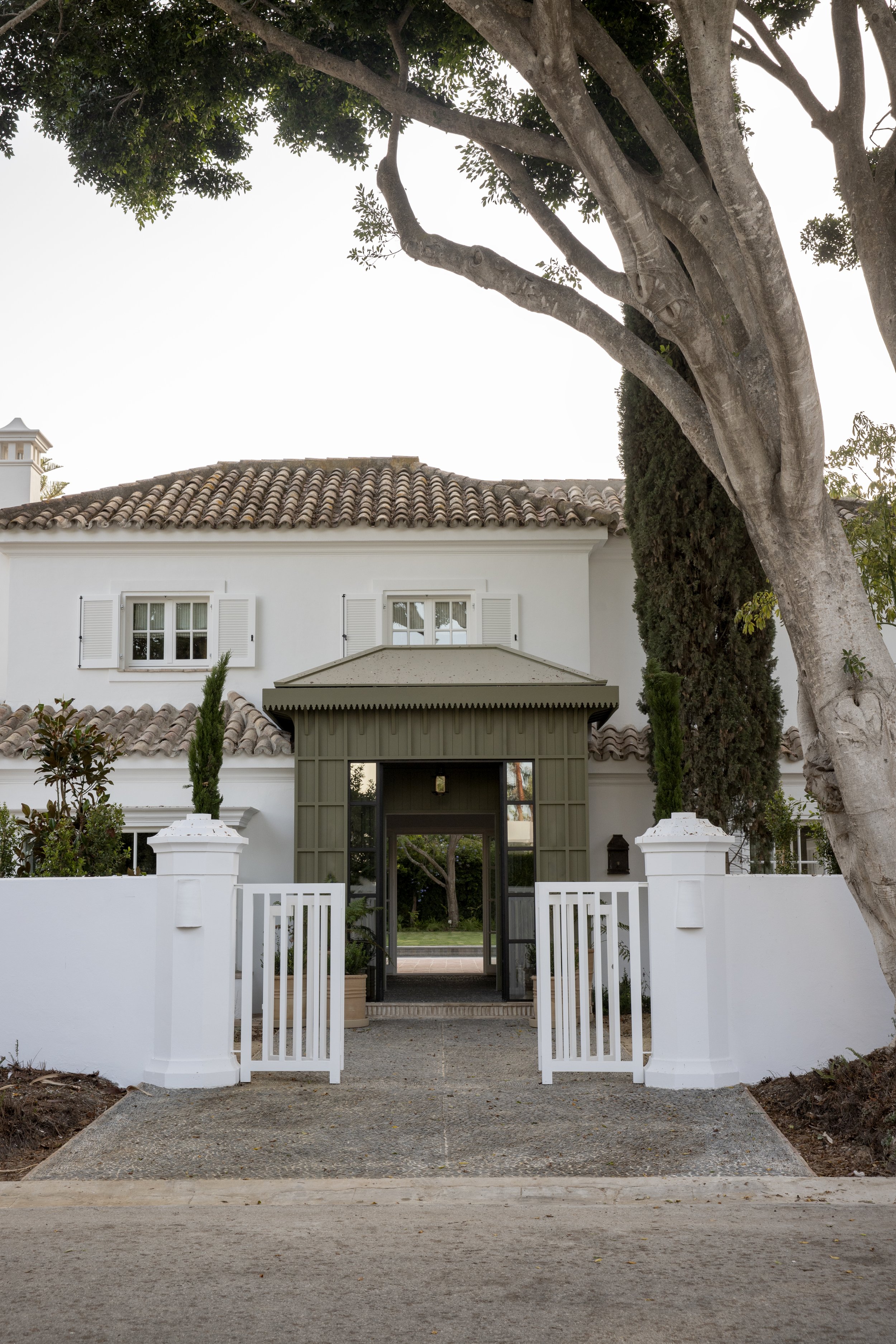 View of a white house with a tiled roof, surrounded by a white fence and open gate, framed by large trees and a garden area.