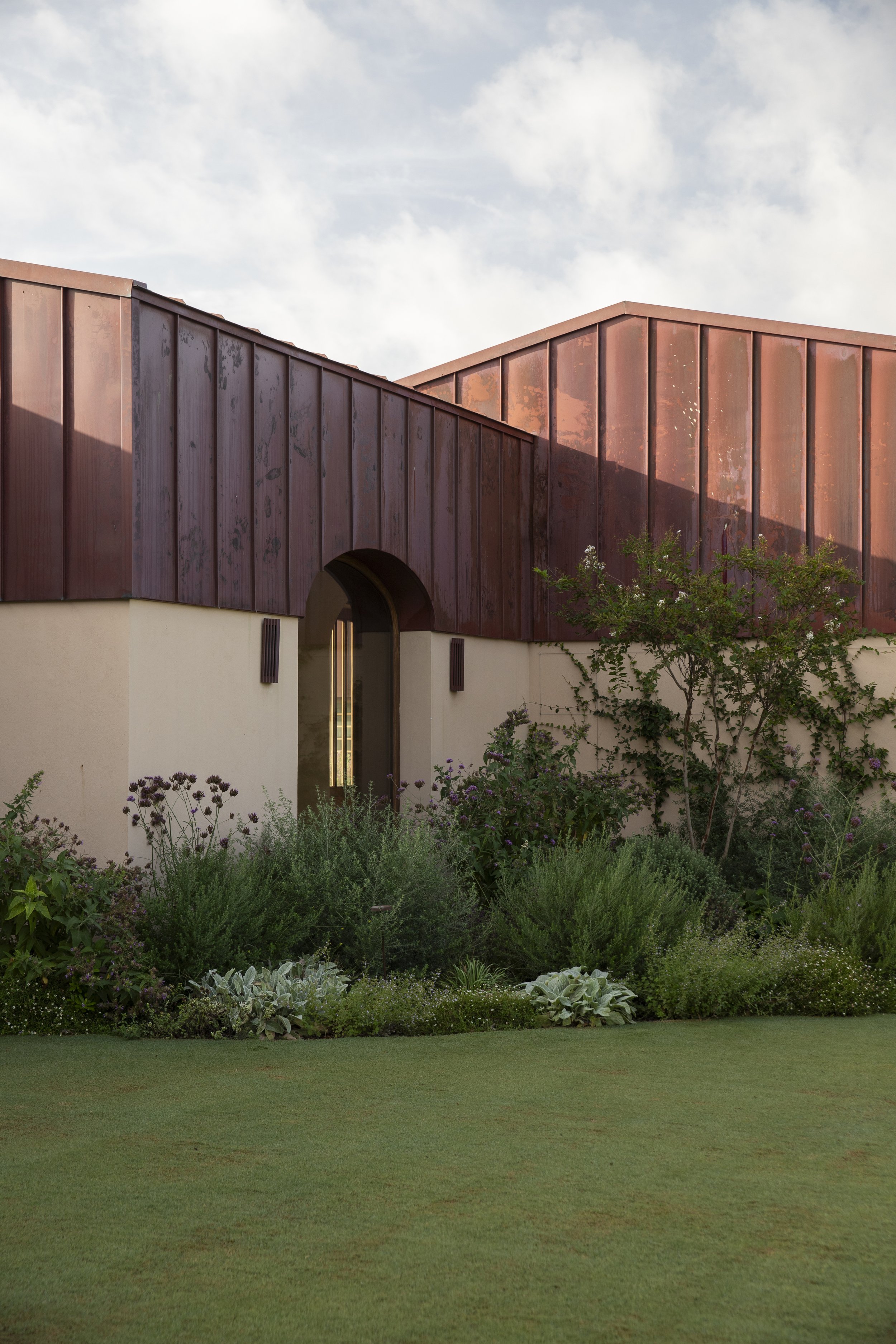 Modern building with reddish-brown metal siding and a small garden in front, under a partly cloudy sky.