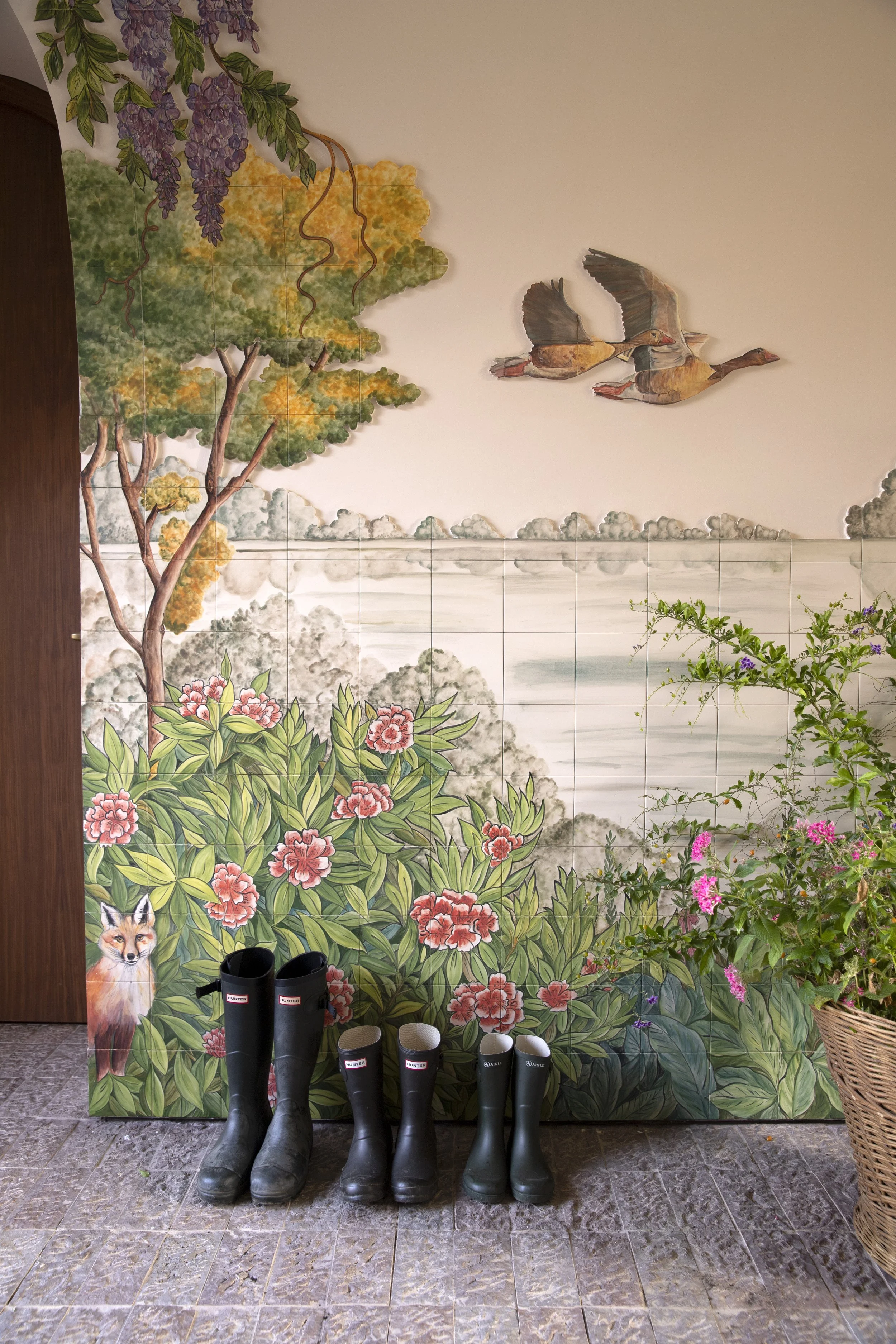 A mural with a landscape and animals, featuring trees, flowering bushes, and two papyrus birds in flight. A fox appears on the lower left corner, and several pairs of black and white boots are lined up in front of the mural on a tiled floor. A wicker