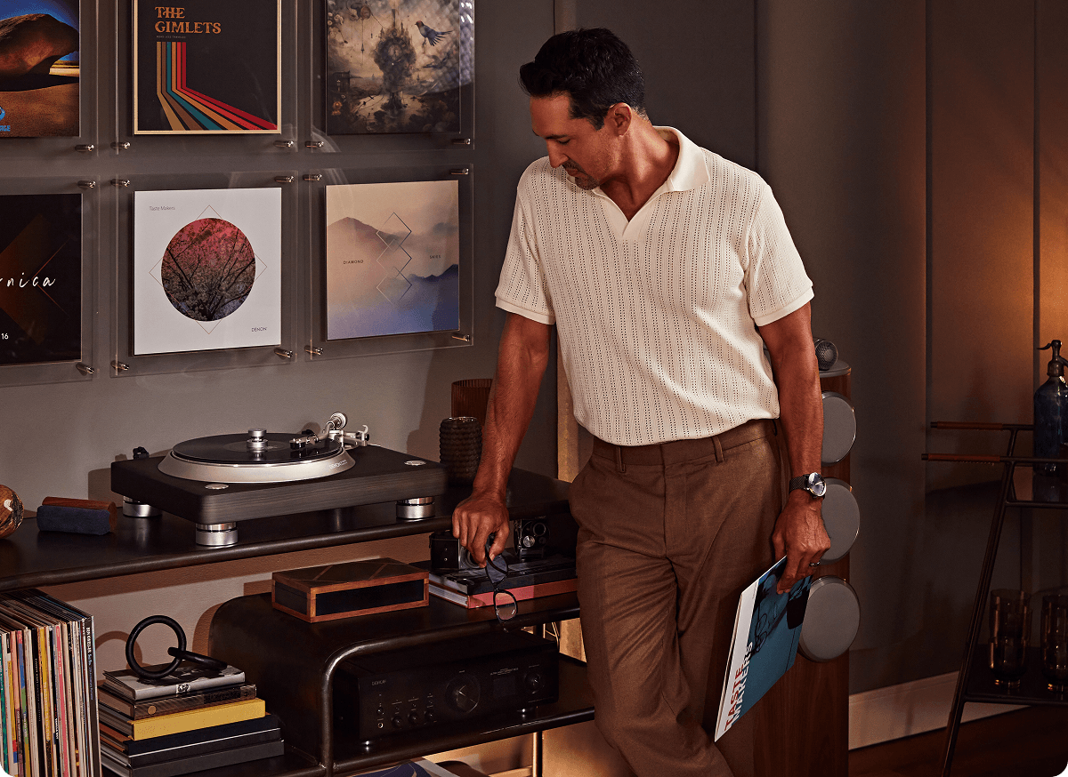 A man adjusts a turntable in a room decorated with framed album covers and wall art, holding a magazine or book in his left hand.