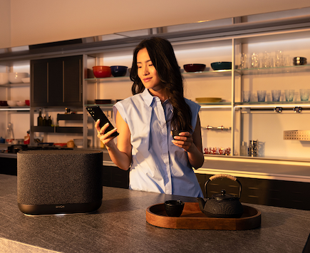 A woman with long dark hair looking at her phone in a modern kitchen, with a speaker and a smart speaker on the countertop.