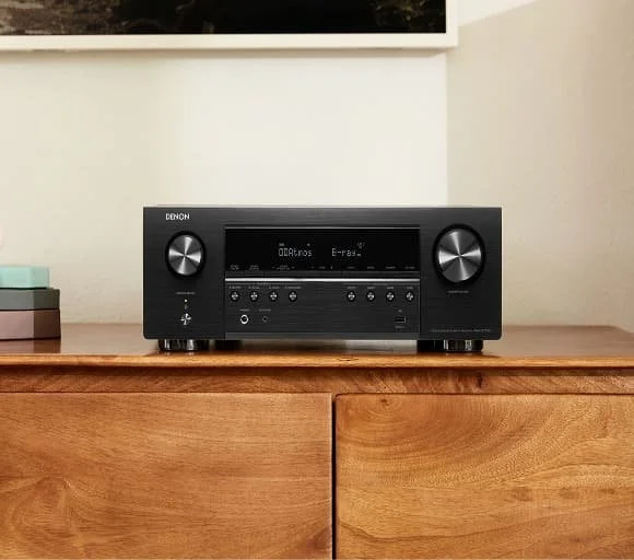 Black Denon audio receiver on a wooden surface with books to the left.