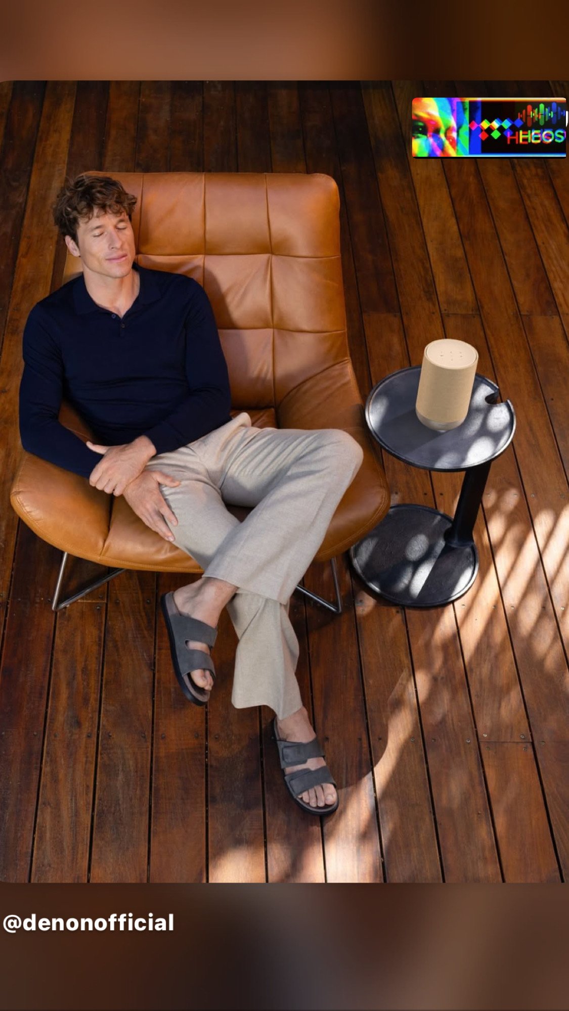 A man with curly hair and light skin sitting in a leather chair with his eyes closed, wearing a dark blue long sleeve shirt, beige pants, and gray sandals in a room with wooden flooring. There is a small round black table beside him with a beige portable speaker on it. A shadow and a pattern of light and shadows are cast on the floor.