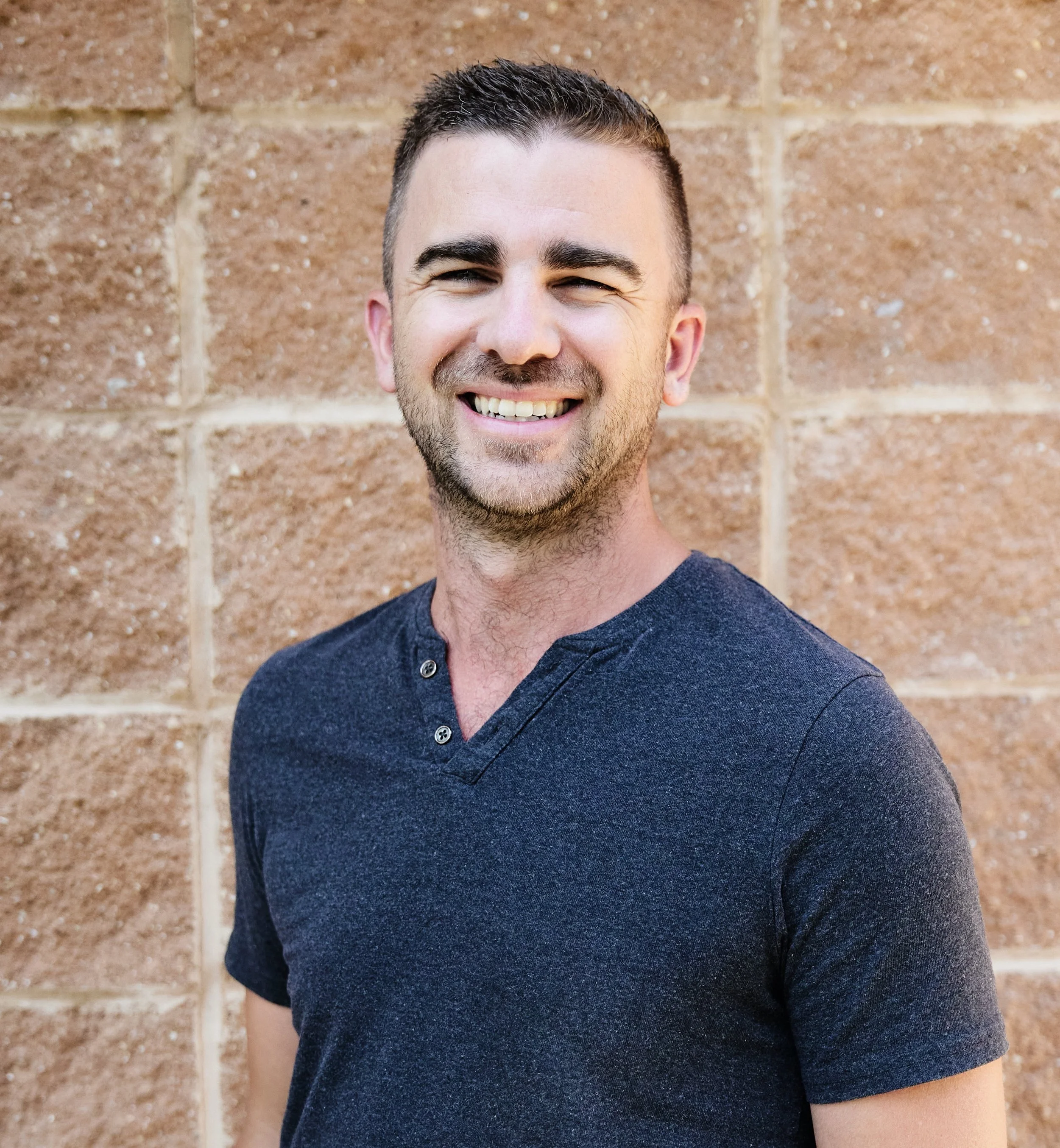 Close-up of a smiling man with short dark hair, slight beard, wearing a dark blue shirt, standing against a brick wall.