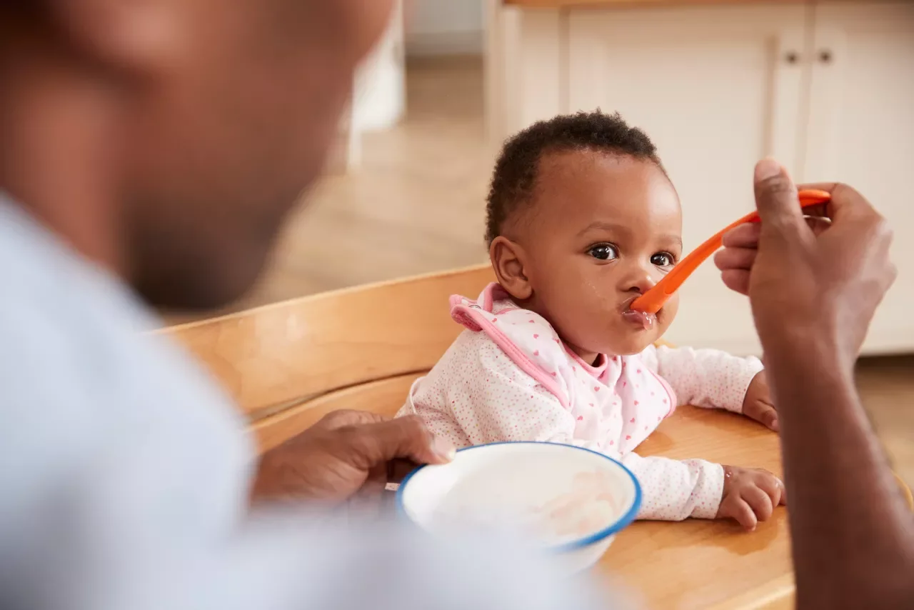A young child with short curly hair sitting in a high chair, being fed with a spoon by an adult. The child looks at the camera as the adult offers food.