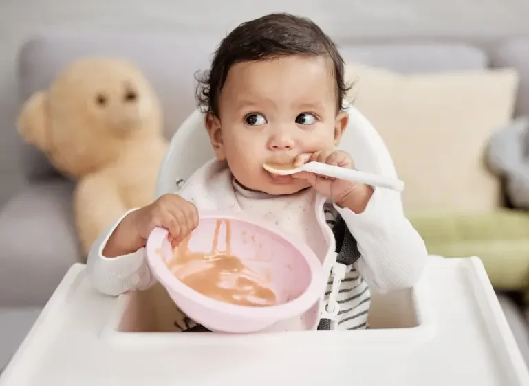 A young child sitting in a high chair, eating with a spoon from a pink bowl, with a teddy bear in the background.