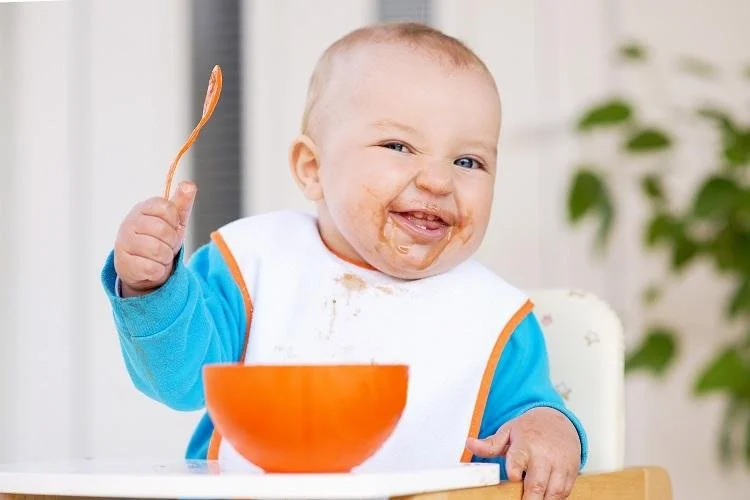 A happy baby with food on their face, holding a spoon in one hand and sitting in a high chair, with an orange bowl in front of them