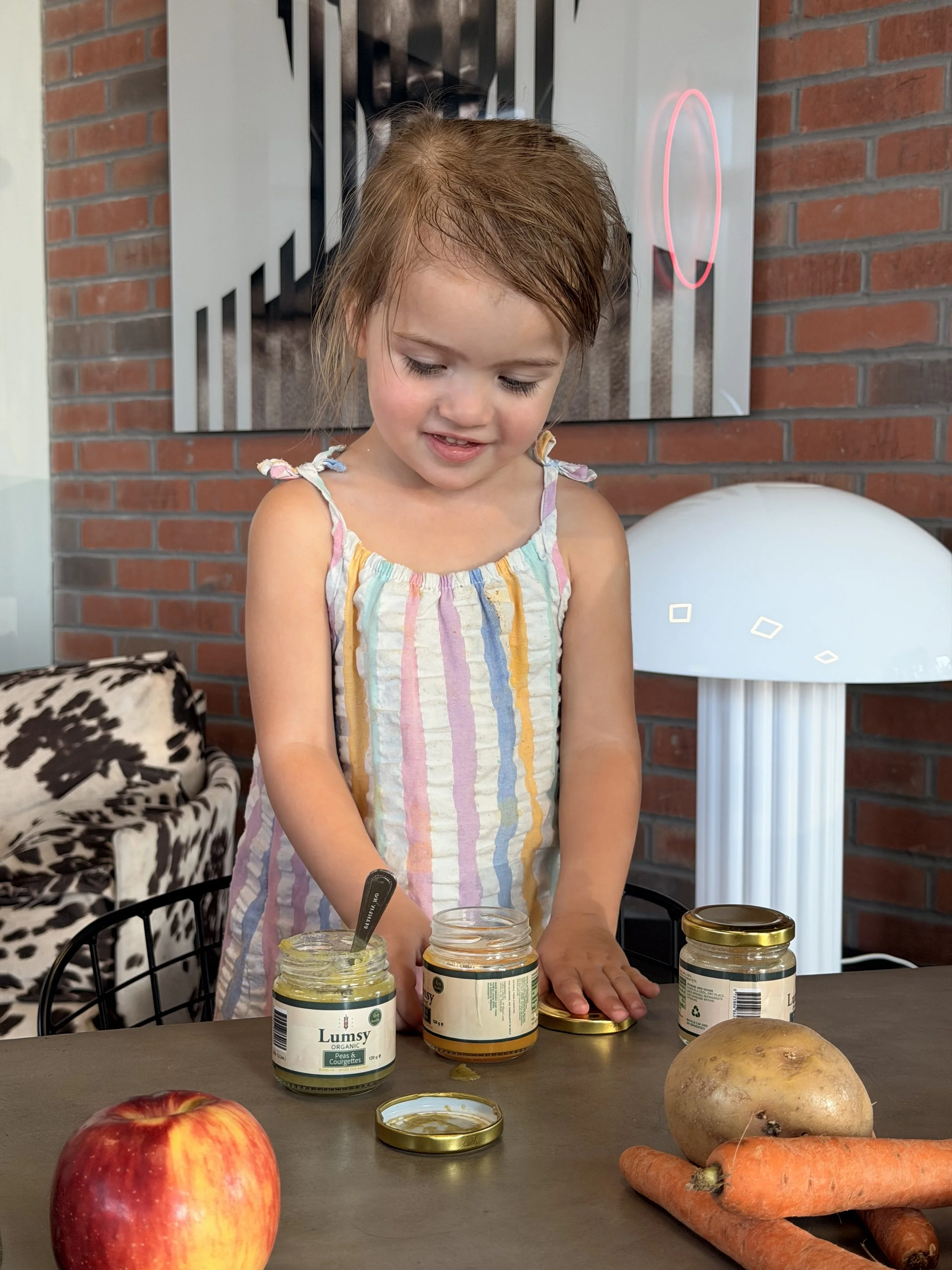 A young girl with light skin and brown hair, wearing a sleeveless, colorful striped dress, is standing at a table with jars of Lumsy organic peas and courgettes. She is looking down and smiling slightly, with her right hand resting on the table. In f