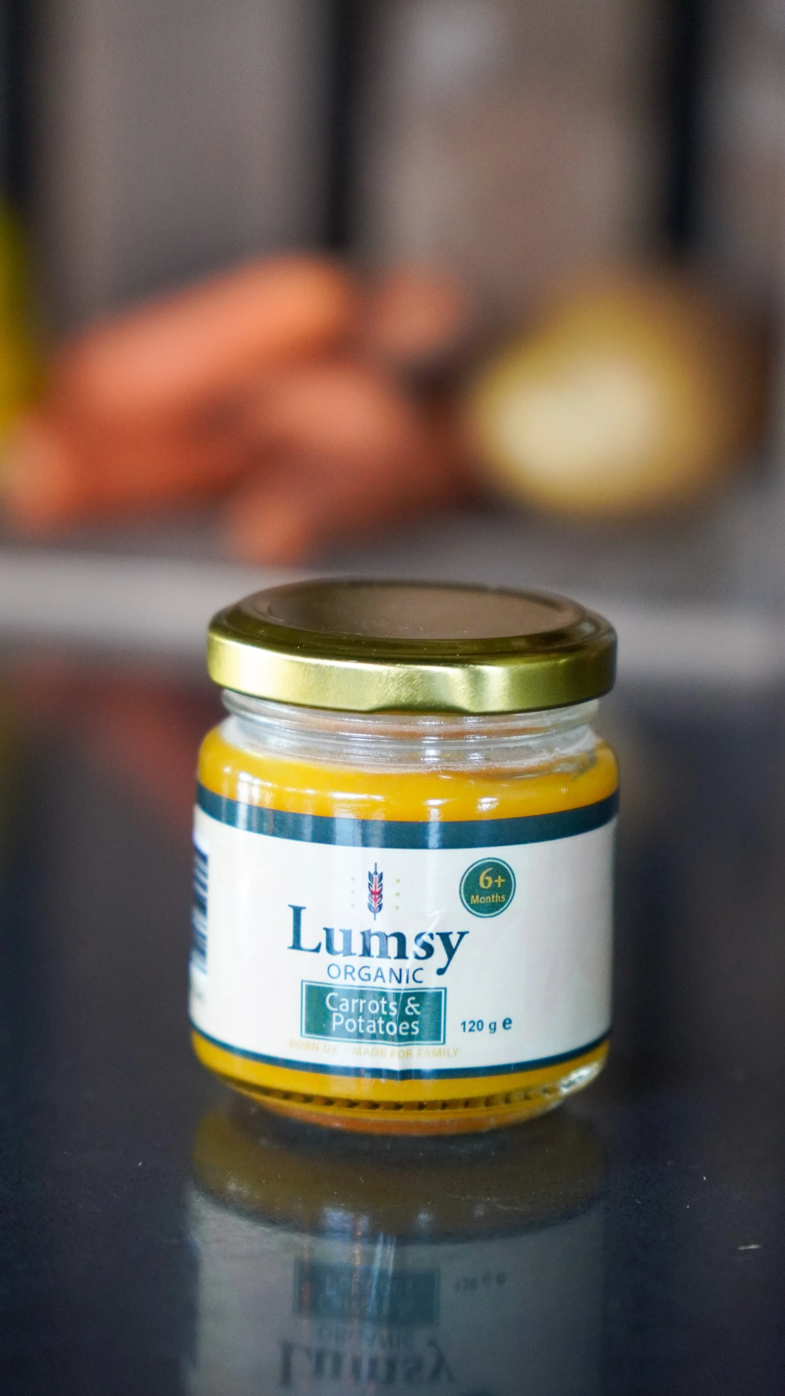 Close-up of a jar of Lumsy Organic Carrots & Potatoes baby food on a reflective surface, with blurred vegetables in the background.