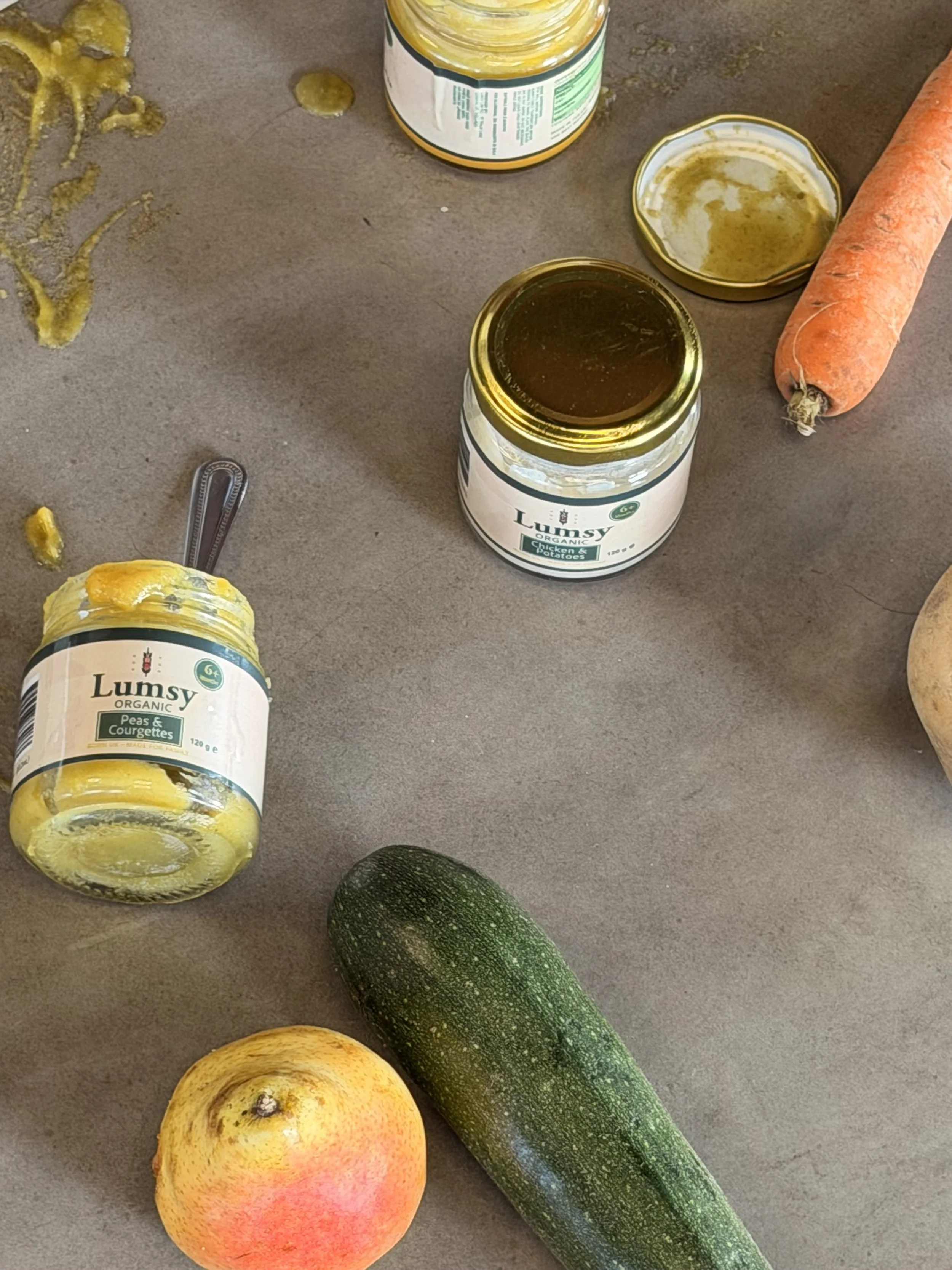 Open jar of creamy peas and courgettes, open jar of pumpkin purée, fresh carrot, whole zucchini, apple, and avocado on a kitchen counter.