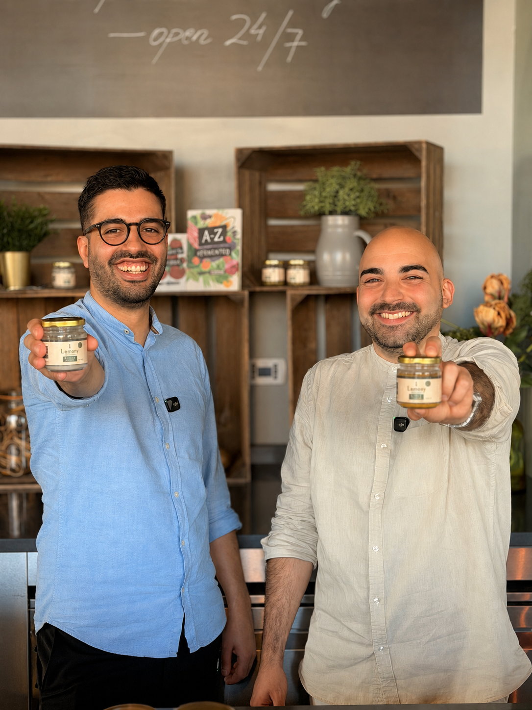 Two smiling men holding jars of lemon spread inside a cafe or shop with wooden shelves, plants, and a chalkboard.