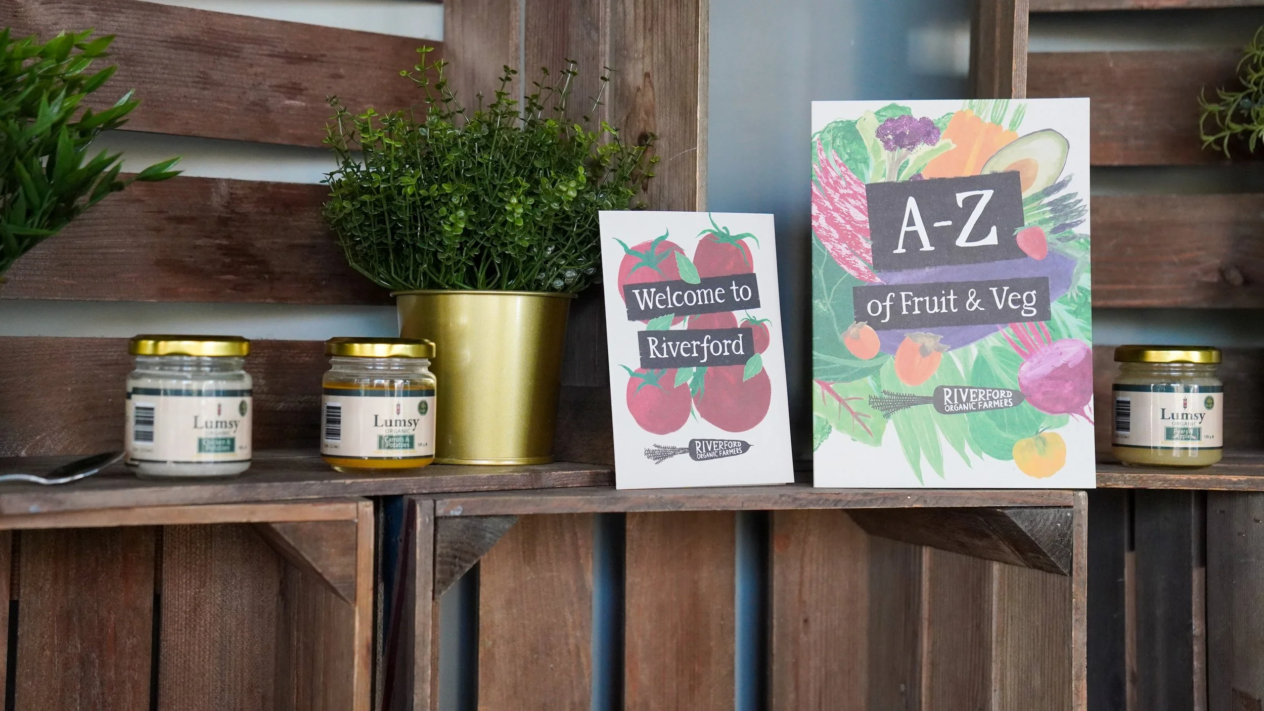 Display of organic farm products including jars of apple and carrot preserves, potted greenery, and colorful signs indicating vegetables and fruits for sale, set against a wooden backdrop.