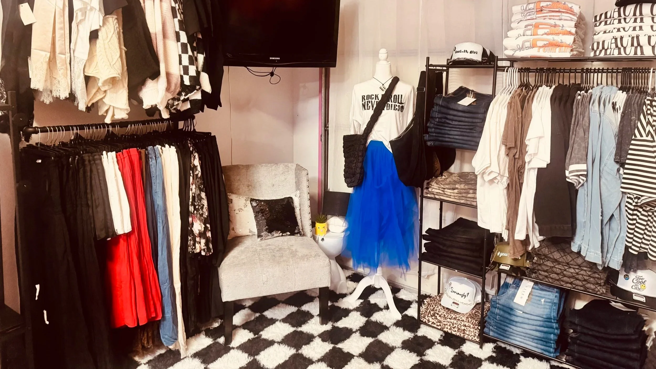 A clothing store fitting room with racks of clothes, a mannequin dressed in a white T-shirt and a blue tulle skirt, and a cozy chair with a pillow, all on a black and white checkered rug.